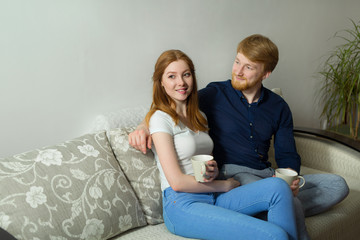 beautiful young couple guy and girl with red hair sitting on the couch and drinking tea