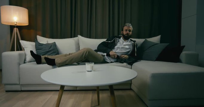 Young adult bearded Caucasian male sits on couch, turning on TV screen. 4K UHD