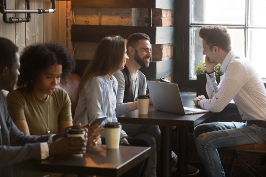 Diverse Multiracial Young People Talking Drinking Coffee Using Devices In Cozy Coffeehouse, Multi-ethnic African And Caucasian Millennials Enjoy Meeting Sitting At Coffeeshop Tables Together In Cafe