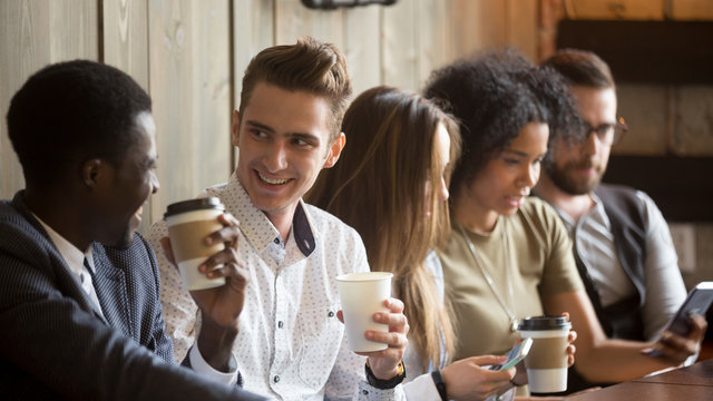 Multiracial young men talking and drinking coffee while other friends using gadgets at meeting in coffeehouse, smiling mates enjoying conversation having fun sitting at cafe table with diverse people