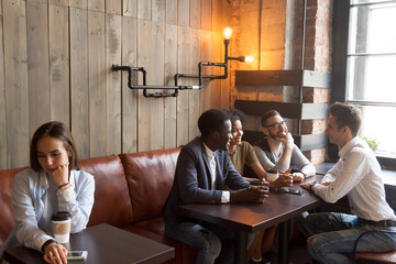Diverse multiracial people hanging together in coffeehouse ignoring sad young girl sitting alone at cafe table, upset social outcast loner suffers from unfair attitude or discrimination among friends - obrazy, fototapety, plakaty