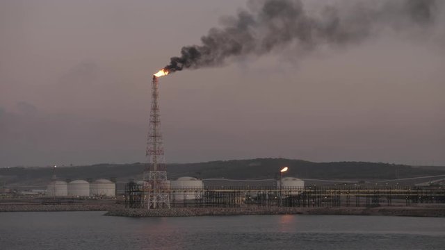 Flare stack with smoke of petrochemical refinery on sea shore in sunset