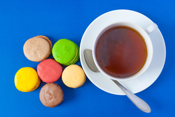 Top view of the french multicolored cakes macarons and a cup of tea on a saucer with spoon on a blue background (flat lay)