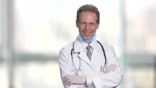 Cheerful handsome physician crossing arms. Male doctor in white suit with folded arms. Abstract blurred windows background.