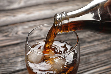 Pouring cola from bottle into glass with ice on wooden background, closeup