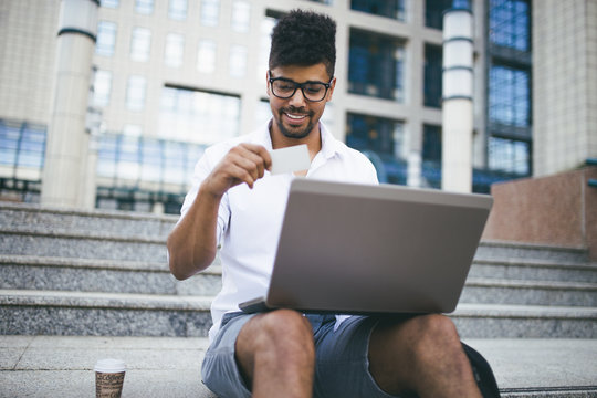 Young attractive fro American businessman sitting on stairs in front of huge modern building and doing some work on laptop