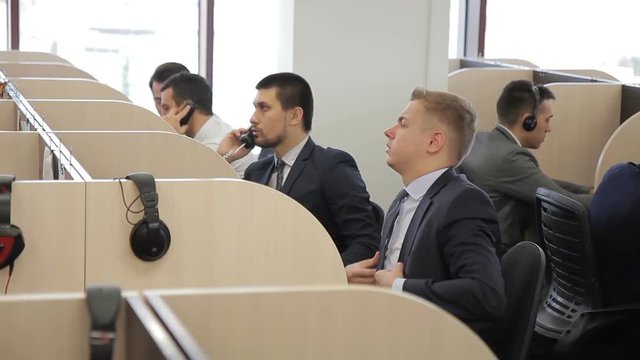 The blone male consultant works among his colleagues in the bright office. The man finishes his phone conversation, eases his tie and starts to call the client. All the workers wear suits and sit in