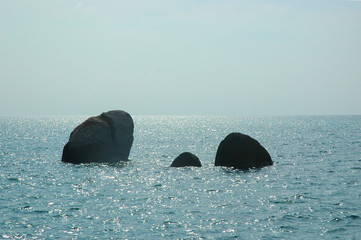 Three dark rocks rising from a sparkling ocean. The sky is pale blue. The ocean stretches to the horizon.