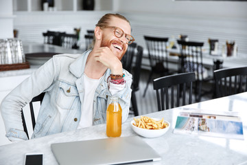 Relaxed cheerful bearded male designer has dinner break after creating project work, enjoys fresh orange juice and french fries, looks happily into distance, sits against spacious cafe interior