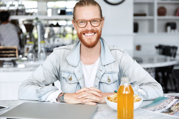 Indoor shot of handsome male with ginger beard, has broad positive smile, rests in cafeteria, eats fast food and drinks juice, surrounded with modern laptop. Stylish smiling male blogger has lunch