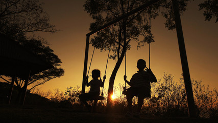 Silhouetted children boy and girl enjoying beautiful sunny day playing on swing set. Red orange...