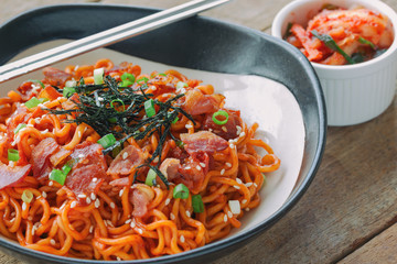 Close up Korean hot and spicy instant noodle in black and white bowl topped with bacon, sesame, seaweed and chopped scallion served with kimchi on wood table. Homemade delicious asian food concept.