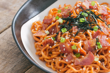 Close up Korean hot and spicy instant noodle in black and white bowl topped with bacon, sesame, seaweed and chopped scallion served with kimchi on wood table. Homemade delicious asian food concept.