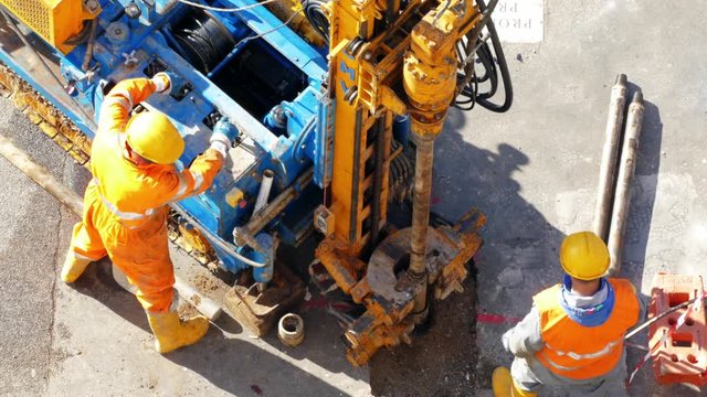 Two men operating powerful metal drilling rig in the centre of the square. Workers using the functional buttons and fixing long metal pipes for taking soil samples