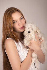 Redhead girl in white dress hugging a dog Maltese at the hands on an isolated grey background