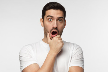 Horizontal portrait of young European man isolated on grey background pictured with eyes wide open with fear or amazement, covering open mouth with palm, astounded with news