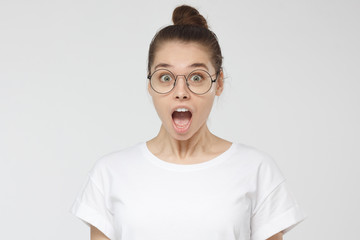 Portrait of positive young woman isolated on grey background wearing thin-rimmed eyeglasses, unable to believe in news concerning surprising details of what she is looking at