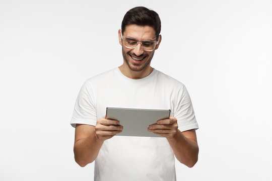 Studio closeup of young man pictured isolated on grey background wearing transparent eyeglasses looking attentively to screen of tablet he is holding, smiling happily, laughing