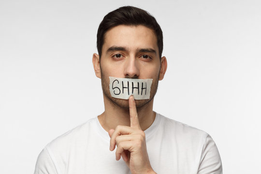 Close up shot of young man with shhh gesture and taped mouth, asking for silence or to be quiet, isolated on grey background