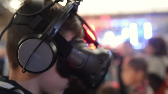 The guy plays in the VR headset virtual reality at a meeting events of computer game fans. In the background there are a lot of people in the blur, bright and lights from concert.