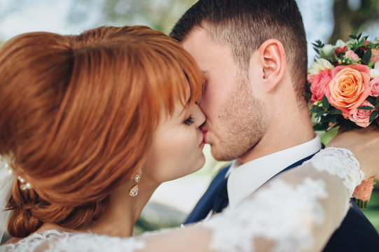 Profile of the kiss of the newlyweds. The red-haired bride. The bride and groom tenderly embrace in the summer park. Loving wedding couple outdoor. Bride and groom.