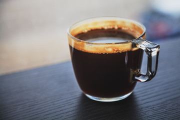 Transparent cup of coffee placed on dark brown laminated wooden surface in front of a window.
