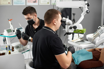 Stomatologist with an assistant in a black gloves treats a patient using a microscope. Modern dentistry with the use of new technologies. Certificates are visible on the wall in the background