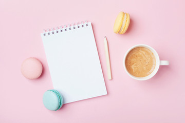 Notebook, cup of coffee and colorful macaron on pastel pink desk top view. Cozy morning breakfast. Fashion flat lay. Sweet macaroons.