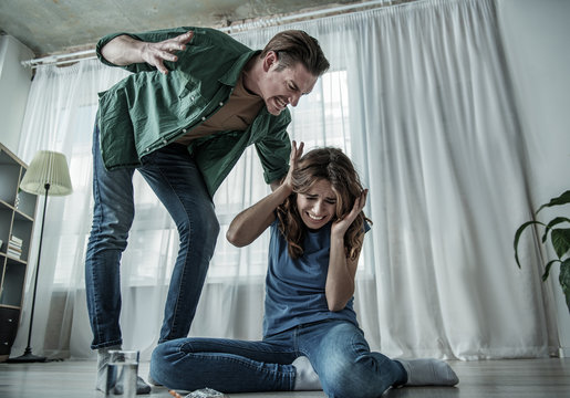 Low angle of irritated husband beating his wife with aggression. Fearful woman is sitting on floor near pills. Family violence concept