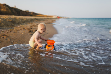 Little cute child baby boy sitting at sea on sand beach, mountain background, play with toy car truck. Little kid son playing outdoors. Family day 15 of may, love, parents, children concept. Side view