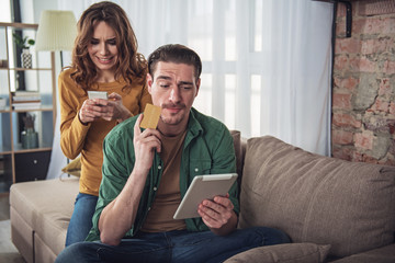 Buy it quickly. Portrait of excited woman using smartphone while doing online shopping at home. Man is holding credit card and looking at tablet thoughtfully