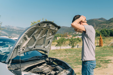 Troubled young man looking under the hood of broken car outdoors in summer. Transportation and...