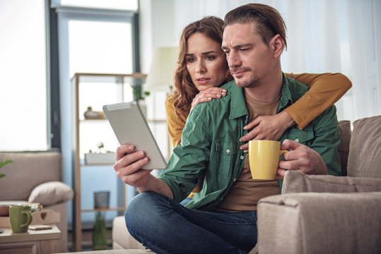 Bad News. Disappointed Man And Woman Looking At Tablet With Amazement. They Are Sitting On Couch At Home