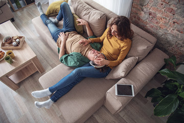 Top view of glad loving couple having rest in apartment. Husband is lying with earphones while wife touching his head with fondness