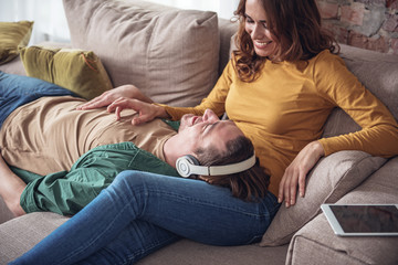 Happy moments together. Carefree married couple relaxing at home in living room. Man is listening to music from headphones while woman is pampering his chest