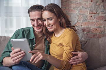 Waist up portrait of happy married couple using tablet with excitement. They are cuddling and...