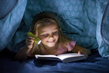 sweet beautiful and pretty little blond girl 6 to 8 years old under bed covers reading book in the dark at night with torch light smiling happy