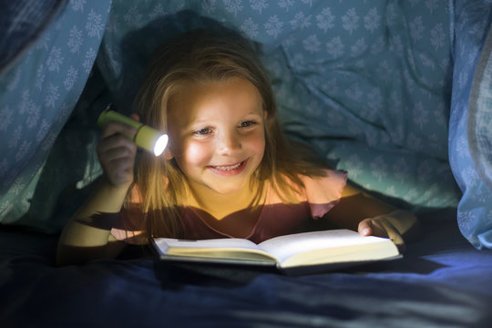 sweet beautiful and pretty little blond girl 6 to 8 years old under bed covers reading book in the dark at night with torch light smiling happy