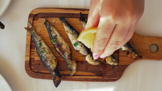 Close-up shot of a woman pouring grilled sardines with lemon juice. Fish served on wooden board