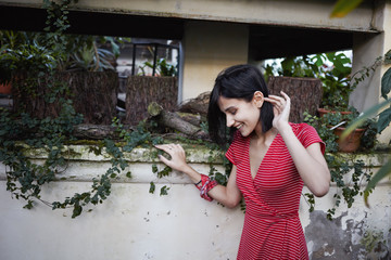 Cheerful young lady wearing red dress with white stripes looking down with cute shy smile, standing isolated at blank wall surrounded by stumps and plants in pots. Nature, greenry and freshness