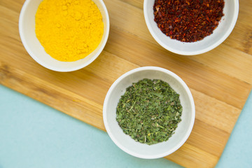 Plastic cups with dry spices and fresh herbs on a wooden cutting board with blue background, top view, close up
