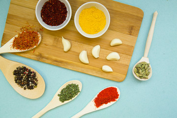 Plastic cups and spoons with dry spices and fresh herbs on a wooden cutting board with blue background, top view, close up