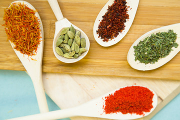 Plastic spoons with dry spices and fresh herbs on a wooden cutting board with blue background, top view, close up