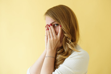 Fototapeta premium Body language. Portrait of fashionable young lady with blonde curly hair making terrified gesture, covering face and peeping through hole between fingers posing in studio against blank wall background