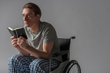 Thoughtful unhealthy man sitting in wheelchair with holy book in hands. Copy space in right side. Isolated on background