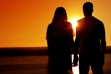 Silhouettes of young romantic couple standing together, holding hands, enjoying summer sunset