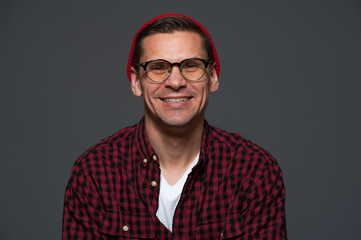 Obraz premium Positive guy hipster in shirt with hat and glasses smiling and posing on gray background