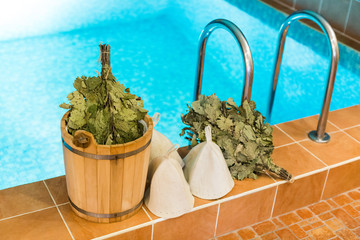 spa, leisure time, healthy life concept. on the edge of swimming pool with clean water there is set for taking bath with fellows that includes few brooms, white wool hats and bucket
