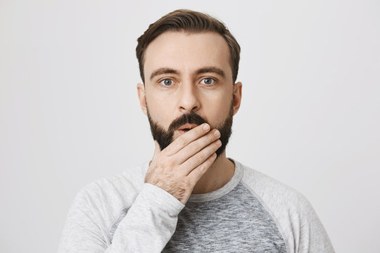 Picture of handsome man with a beard expressing surprise covering his mouth with hand over white background. Coworker just told an interesting gossip about their employer.