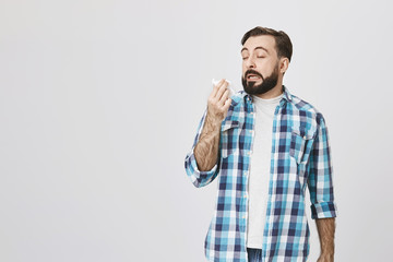 Studio shot of sick man who is about to sneeze with closed eyes and opened mouth while holding napkin in hand, standing over gray background. Friend has cat and this guy is allegic to it.